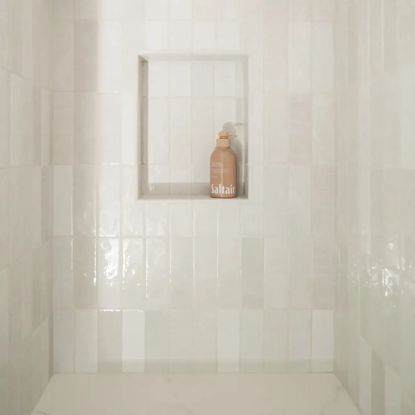 Bright white ceramic tile shower with a recessed shelf displaying a bottle of Saltair.