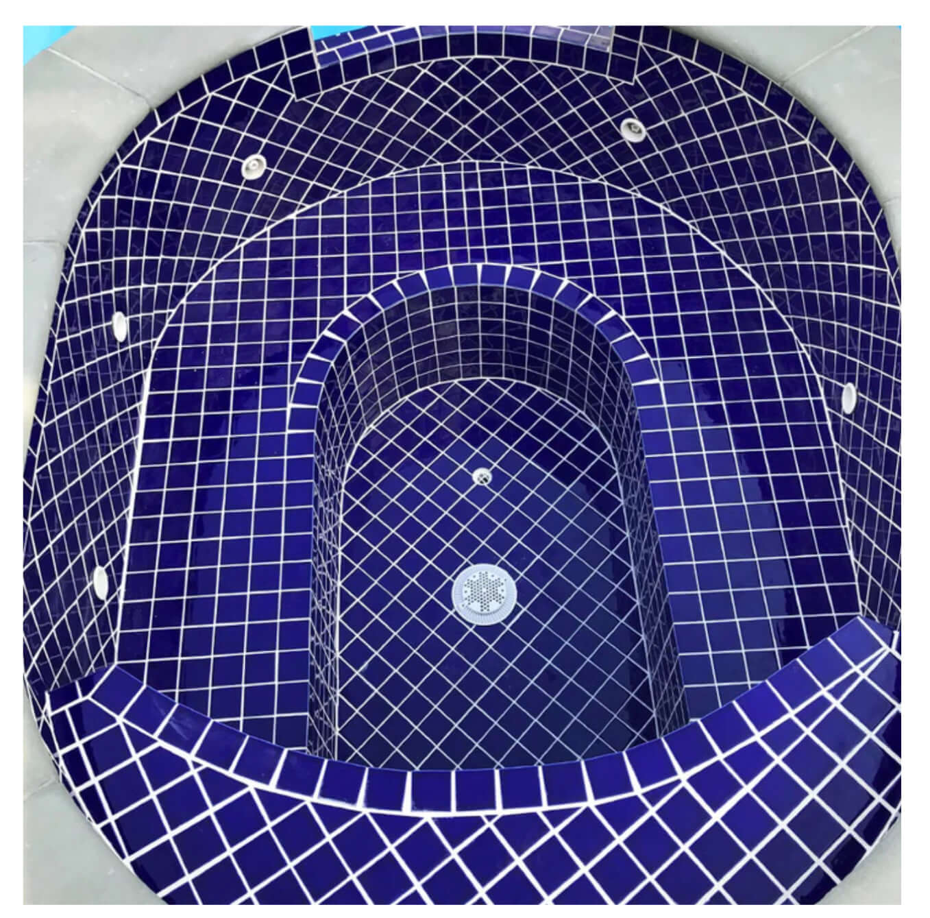 Top view of a cobalt blue mosaic tiled hot tub, featuring a sleek oval design with modern plumbing fixtures.