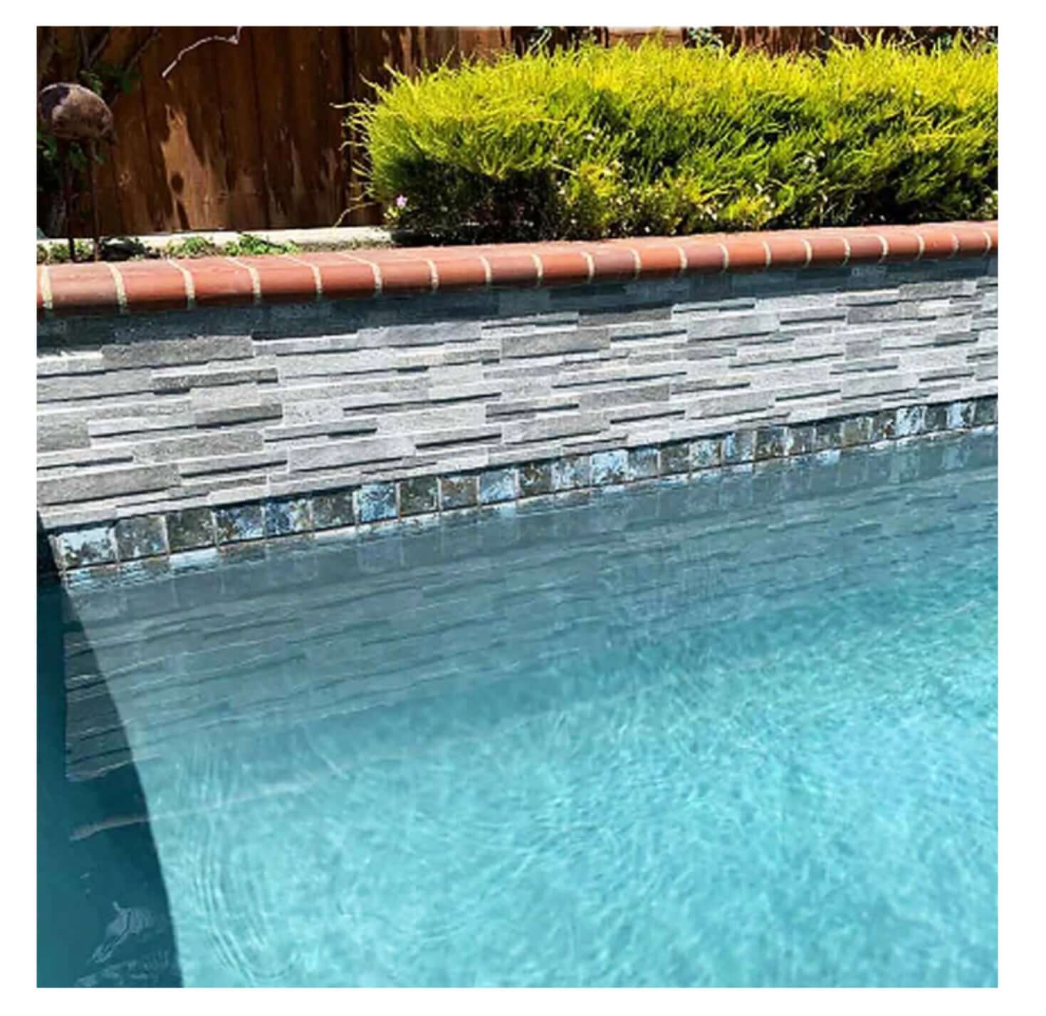 Close-up of a pool wall featuring gray stacked stone tiles and a brick border, surrounded by lush greenery.