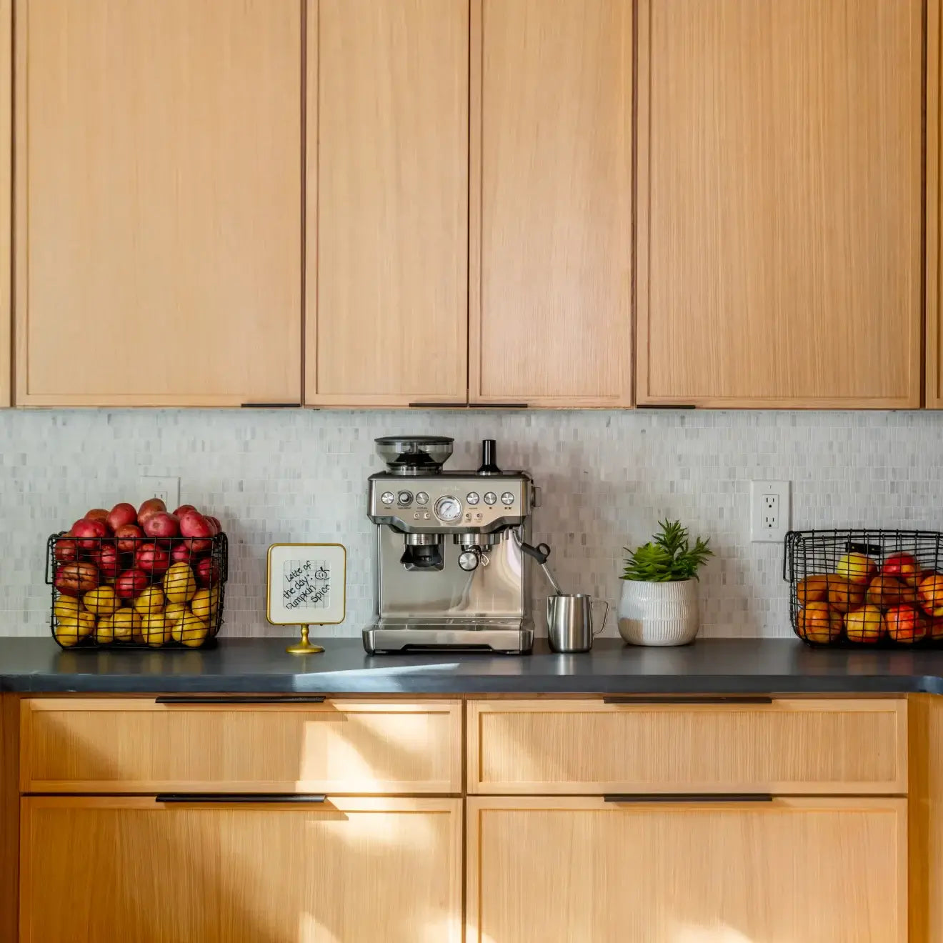 Modern kitchen with wooden cabinets, espresso machine, and baskets of colorful apples on the countertop.